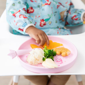 A child in a high chair enjoys a meal from Bumkins Silicone Plate + Spoon Set: Disney Princess with broccoli, mashed potatoes, and nuggets.