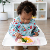 A toddler in a bib sits in a high chair, eating from Bumkins Silicone Plate + Spoon Set: Disney Princess with veggies, nuggets, and potatoes.