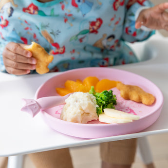 A child in a colorful bib enjoys nuggets, mashed potatoes, and fruit from Bumkins Silicone Plate + Spoon Set: Disney Princess.