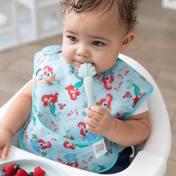 A baby in a mermaid bib sits in a high chair, holding a spoon from Bumkins Silicone Plate + Spoon Set: Disney Princess Ariel.