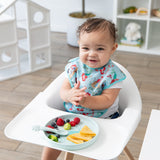 A happy baby in a high chair and colorful bib enjoys snacks from Bumkins Silicone Plate + Spoon Set: Disney Princess Ariel.