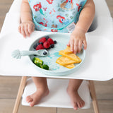 A baby in a high chair wearing a bib eagerly reaches for food from Bumkins Silicone Plate + Spoon Set: Disney Princess Ariel.