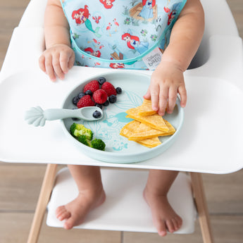 A baby in a high chair wearing a bib eagerly reaches for food from Bumkins Silicone Plate + Spoon Set: Disney Princess Ariel.