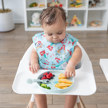 A baby sits in a high chair wearing a bib, eagerly reaching for food from Bumkins Silicone Plate + Spoon Set: Disney Princess Ariel.