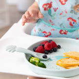 A child reaches for food in a high chair from Bumkins Silicone Plate + Spoon Set: Disney Princess Ariel, wearing a mermaid-print bib.