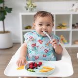A baby in a high chair eats from Bumkins Silicone Plate + Spoon Set: Disney Princess Ariel, adorned with fruit slices and berries.