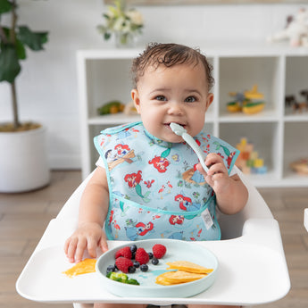 A baby in a high chair eats from Bumkins Silicone Plate + Spoon Set: Disney Princess Ariel, adorned with fruit slices and berries.