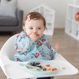 A girl wearing a blue bib smiles as she eats from a Bumkins Silicone Plate + Spoon Set: Disney Princess Ariel in a high chair.