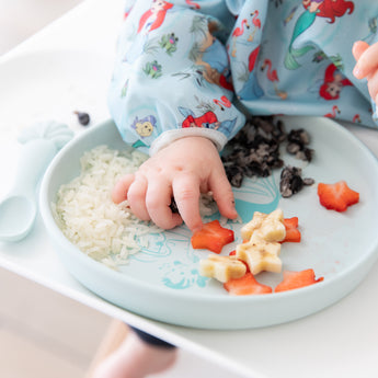 Close-up of a child in a blue bib eating fruit, rice, & beans from Bumkins Silicone Plate + Spoon Set: Disney Princess Ariel.