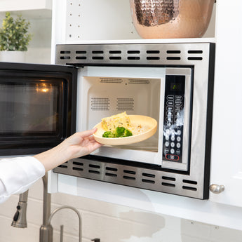 A person places Bumkins Silicone Plate + Spoon Set: Disney Winnie the Pooh filled with food into an open microwave.