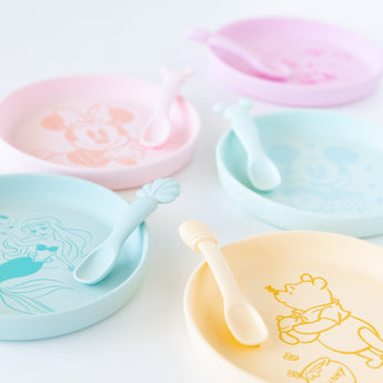 Bumkins Silicone Plate + Spoon Set: Disney Winnie the Pooh sits on a white background next to other similar plate sets.