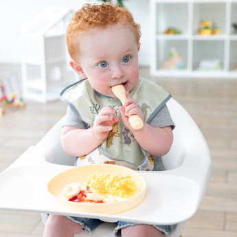A curly red-haired baby sits in a high chair, holding a spoon with Bumkins Silicone Plate + Spoon Set: Disney Winnie the Pooh on the tray.