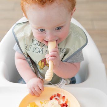 A red-haired baby in a high chair with a bib enjoys bananas and strawberries from Bumkins Silicone Plate + Spoon Set: Disney Winnie the Pooh.