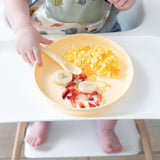 A toddler enjoys breakfast from a Bumkins Silicone Plate + Spoon Set: Disney Winnie the Pooh on a high chair's white tray.