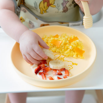 A child wearing a bib in a high chair enjoys breakfast from a Bumkins Silicone Plate + Spoon Set: Disney Winnie the Pooh.