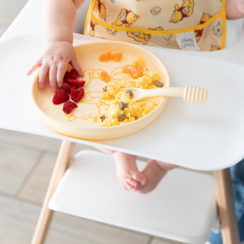 A baby in a high chair reaches for raspberries from Bumkins Silicone Plate + Spoon Set: Disney Winnie the Pooh beside eggs and oranges.
