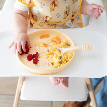 A baby in a high chair enjoys eggs and raspberries from a Bumkins Silicone Plate + Spoon Set: Disney Winnie the Pooh while wearing a bib.