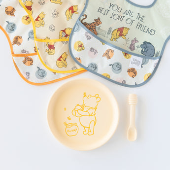 Bumkins Silicone Plate + Spoon Set: Disney Winnie the Pooh lays on a white background next to three waterproof bibs.