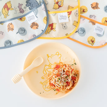 Bumkins Silicone Plate + Spoon Set: Disney Winnie the Pooh, filled with pasta, is displayed next to bibs on a white background.