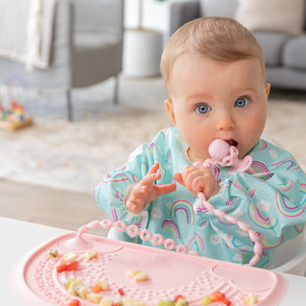 A bright-eyed baby chews a pink toddler fork attached to a Bumkins Silicone Accessory Tether 2-Pack: Pink on a pink placemat with fruit.