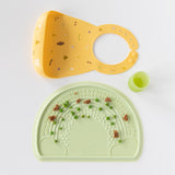 Bumkins Silicone Bib: Camp Gear rests above a green cup and green placemat with peas and bits of food on a white background.