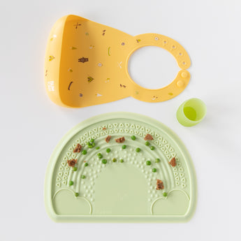 Bumkins Silicone Bib: Camp Gear rests above a green cup and green placemat with peas and bits of food on a white background.