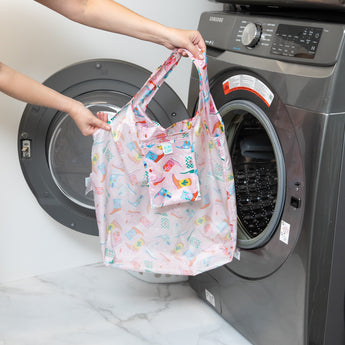 A person holds a bag from Bumkins The Playdate Planner Deluxe 5-Piece Set: Boots in front of an open front-loading washing machine.