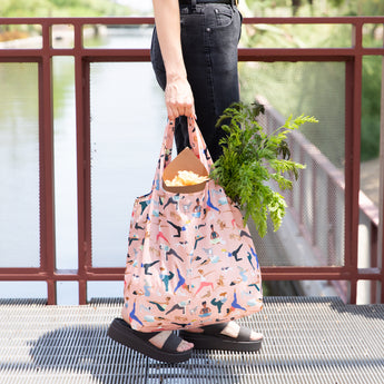 A person holding a Bumkins Packable Tote Bag: Yoga Pals with groceries, including greens, stands on a metal bridge.