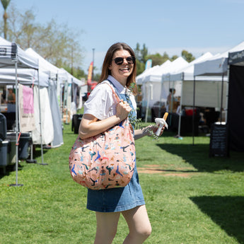 A smiling woman at outdoor market with Bumkins Packable Tote Bag: Yoga Pals on her shoulder holds a snack, wearing sunglasses and denim dress.