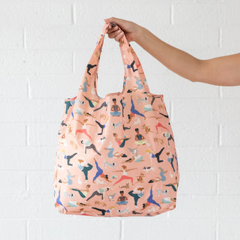 A hand holds a Bumkins Packable Tote Bag: Yoga Pals against a white brick wall.