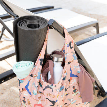 Bumkins Packable Tote Bag: Yoga Pals, made of waterproof fabric, hangs on a chair by the pool, holding a yoga mat, bottle, and towel.