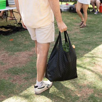 A person in shorts and sneakers holding a Bumkins Packable Tote Bag: Rich Black with greens at an outdoor market.