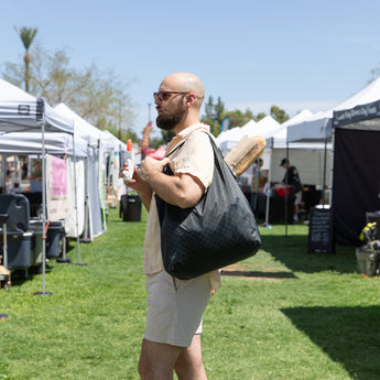 A man with a Bumkins Packable Tote Bag: Rich Black, baguettes, and iced coffee strolling through a sunny outdoor market with white tents.