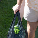 Close-up of a person in shorts holding a Bumkins Packable Tote Bag: Rich Black, with fresh broccoli visible. Grass is seen in the background.