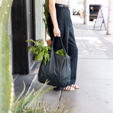 A person on a sidewalk holding a Bumkins Packable Tote Bag: Rich Black filled with fresh green leafy veggies.