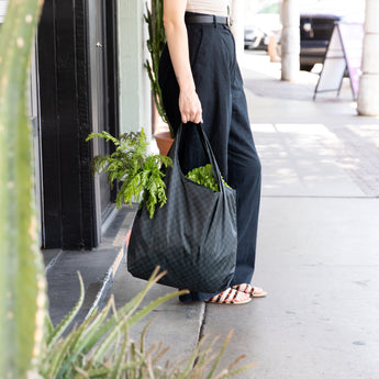 A person on a sidewalk holding a Bumkins Packable Tote Bag: Rich Black filled with fresh green leafy veggies.