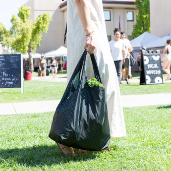 At an outdoor market, a person in a white dress carries a Bumkins Packable Tote Bag: Rich Black with greenery peeking out.