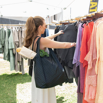 A woman shops at an outdoor market, carrying a Bumkins Packable Tote Bag: Rich Black with a baguette and greens.