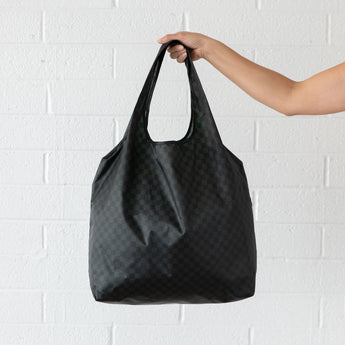 A hand holds a Bumkins Packable Tote Bag: Rich Black against a white brick wall.