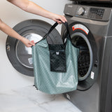 A person holds a Bumkins Packable Tote Bag: Rich Black in front of an open front-loading washing machine.