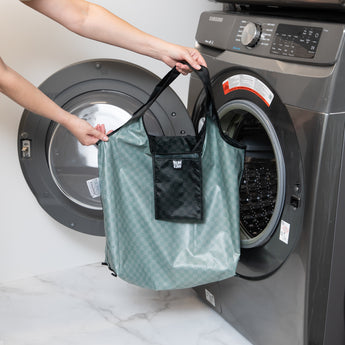 A person holds a Bumkins Packable Tote Bag: Rich Black in front of an open front-loading washing machine.