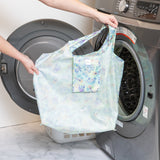 A person holds a bag from Bumkins The Playdate Planner Deluxe 5-Piece Set: Aloha Stitch in front of an open front-loading washing machine.