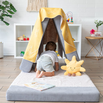 A toddler in a white shirt and light shorts crawls into a pillow fort with a Bumkins Reversible Roll & Go Blanket, Desert Boho on top.