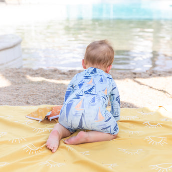 A baby in a blue sailboat onesie sits on a Bumkins Reversible Roll & Go Blanket, Desert Boho on the sandy beach by sparkling water.