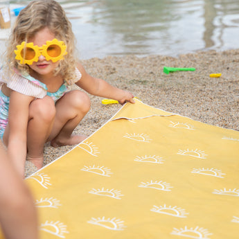 A child in sun-shaped sunglasses arranges a Bumkins Reversible Roll & Go Blanket, Desert Boho on a pebbly beach with sand toys nearby.