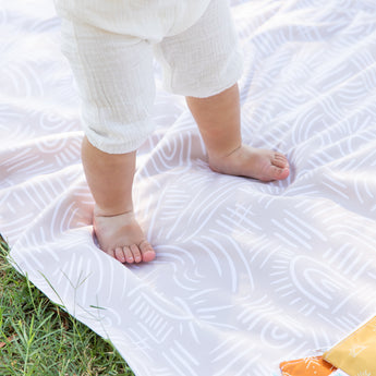 Close-up of a toddler in white pants standing on a Bumkins Reversible Roll & Go Blanket, Desert Boho outside, with green grass visible.