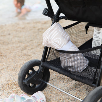 A stroller with a rolled Bumkins Reversible Roll & Go Blanket, Desert Boho is parked on a sandy beach. A child plays in the background.