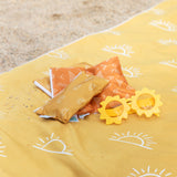 Sun-shaped sunglasses and small bags atop a Bumkins Reversible Roll & Go Blanket, Desert Boho on the beach.