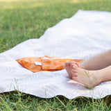 A child's feet rest on a Bumkins Reversible Roll & Go Blanket, Desert Boho, spread over the grass with small orange bags nearby.