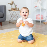 A toddler kneels on a Bumkins Reversible Roll & Go Blanket, Desert Boho, holding teething rings in a bright living room.